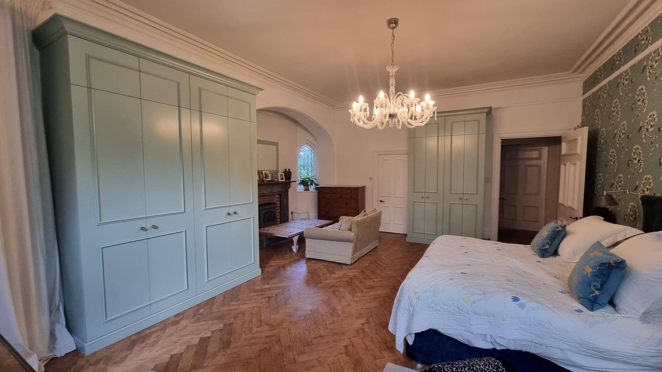 Two sets of custom fitted wardrobes in sage green framing a large period bedroom with herringbone flooring