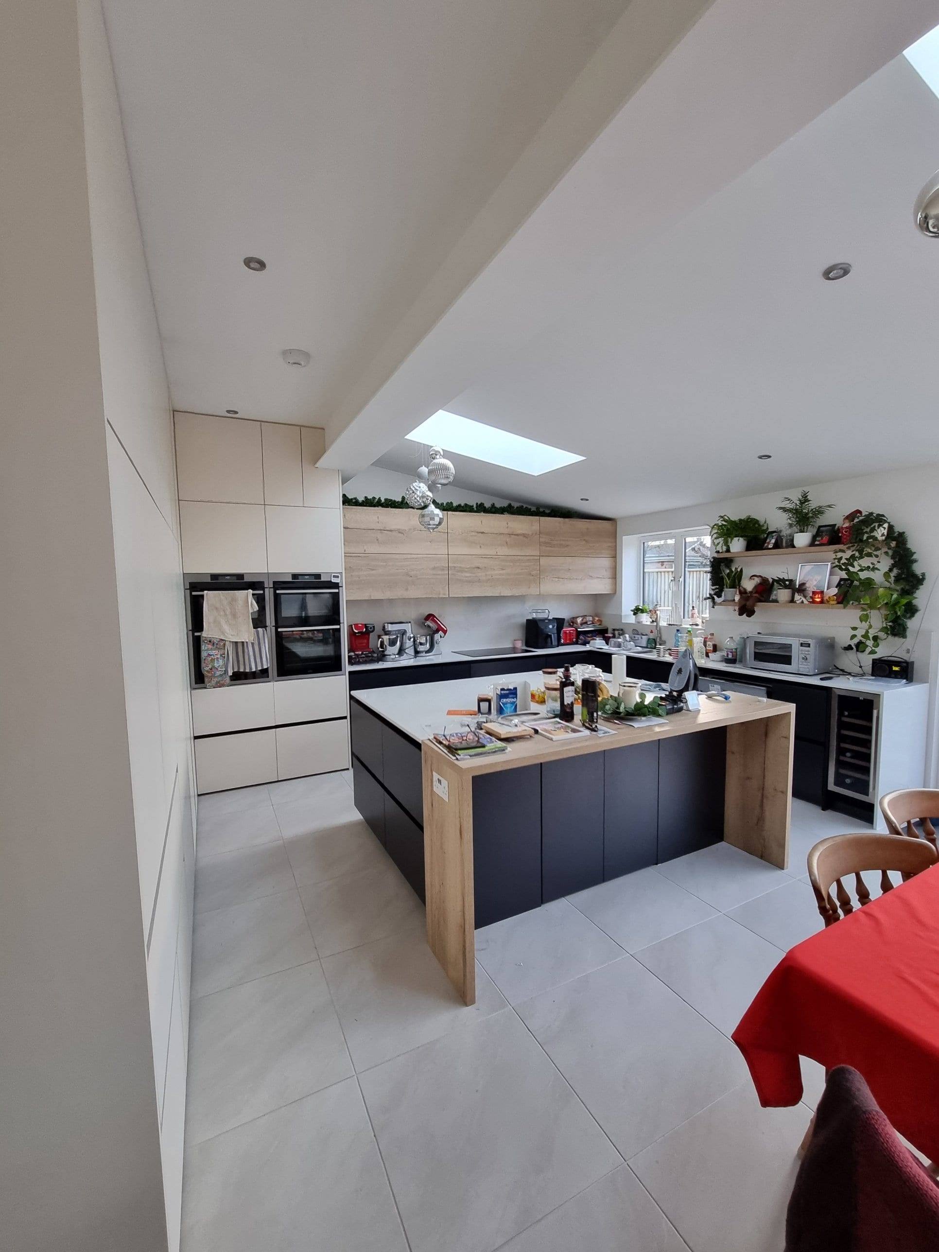 Open-plan handleless kitchen with a dark island, oak waterfall end panel, tall cream units and a rooflight above