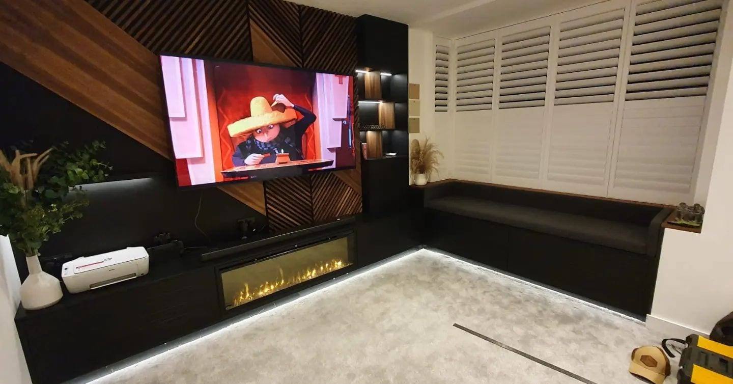 Evening view of a fitted media wall with an inset electric fire, window seat storage and LED plinth lighting