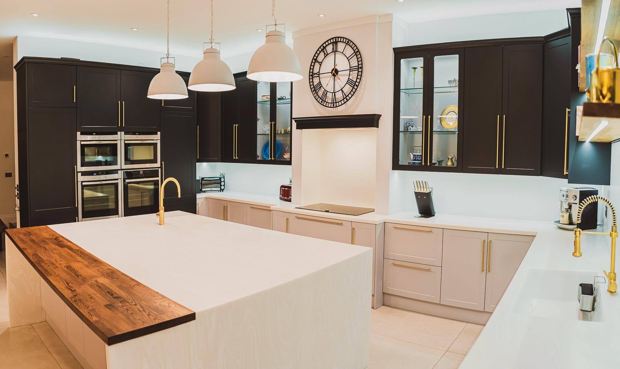 Custom painted shaker kitchen with glazed display cabinets, brass bar handles, integrated ovens and a feature clock above the hob